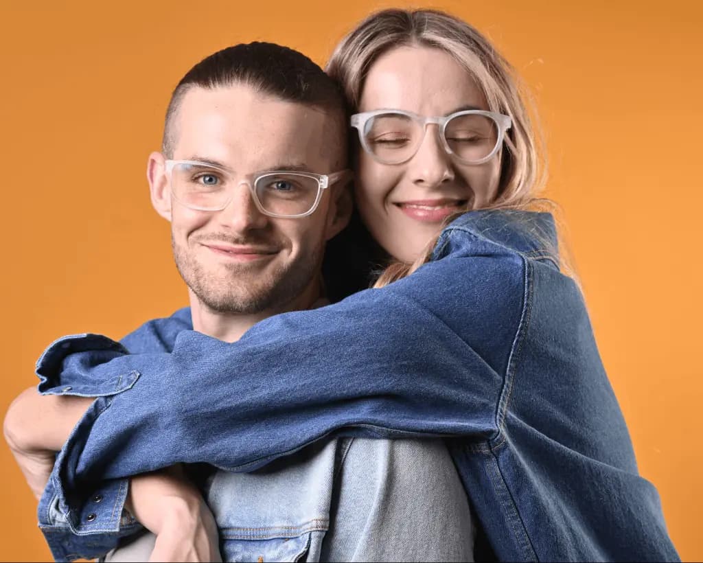 Two people wearing modern Dresden Vision eyeglasses share a warm embrace against an orange background, both smiling and wearing denim