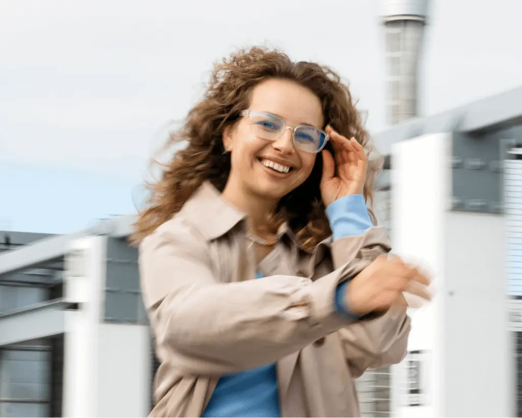 A woman in a beige jacket laughing outdoors while adjusting her Dresden Vision glasses, with modern architecture in the background