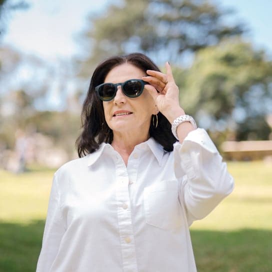 Stylish woman wearing Polarised Sunglasses in New Zealand, smiling in natural sunlight, dressed in a white blouse with hoop earrings, relaxing in a lush green public park.