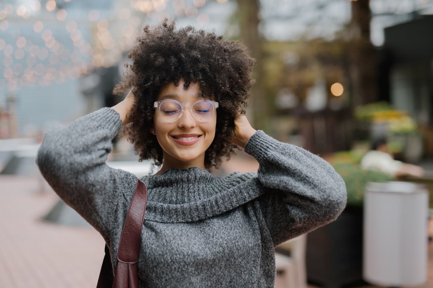 Young woman wearing stylish bifocal lenses in Canada, smiling with eyes closed, enjoying crisp outdoor air in a cosy sweater.