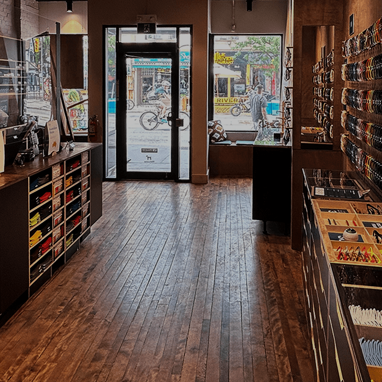 Interior view of the Dresden Vision store in Little Italy, Toronto, featuring rows of colorful eyewear frames on wooden wall displays, a sleek dark wood service counter with organized drawers, and warm hardwood floors under bright, modern lighting.