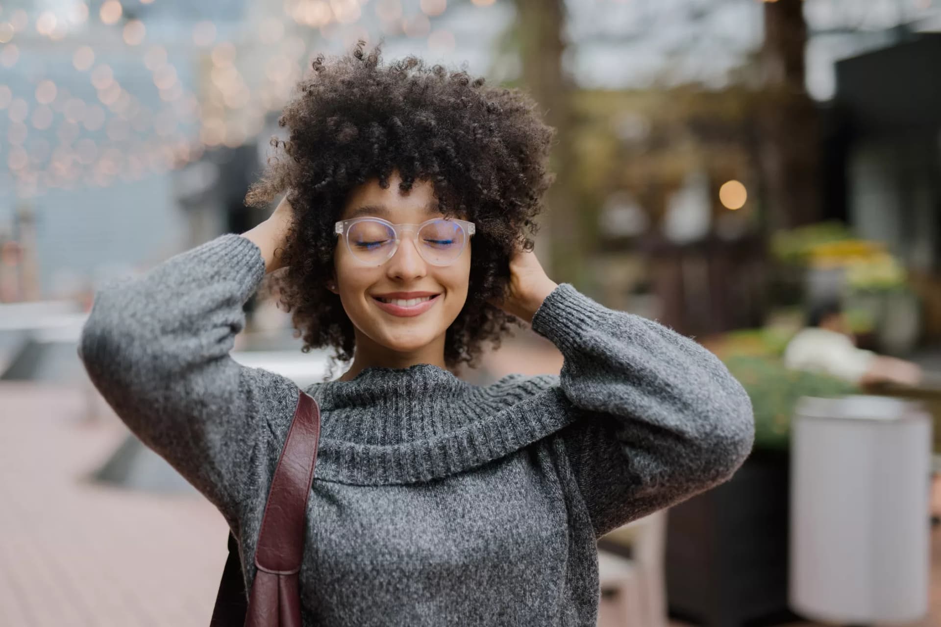 Smiling woman in clear bifocal lenses in Australia wearing a grey sweater outdoors with her hands in curly hair.